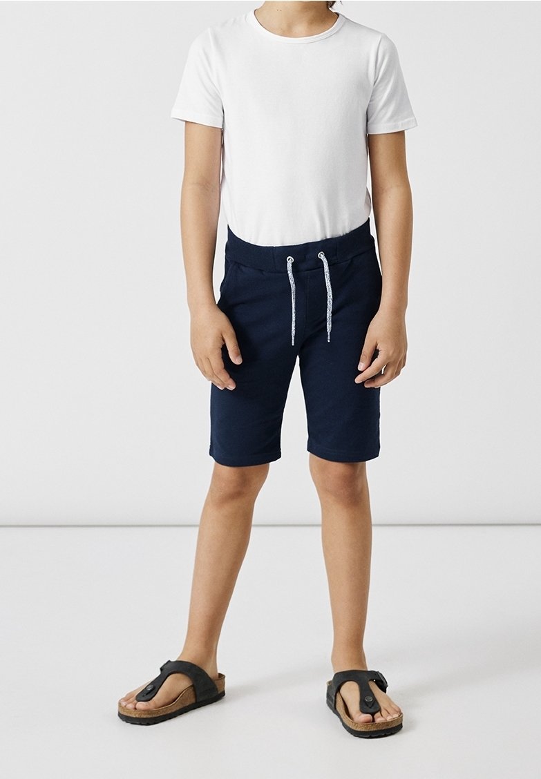 Enfant portant une chemise blanche à manches courtes, un short bleu marine avec cordon de serrage, et des sandales noires à bout ouvert, debout sur un fond blanc uni.