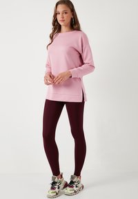 Felpa rosa a maniche lunghe con vestibilità rilassata e fessure laterali; abbinata a leggings bordeaux e scarpe da ginnastica multicolori.
