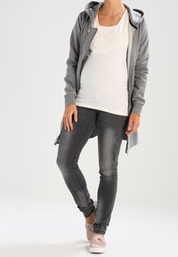 Felpa grigia con zip e tasche laterali, top bianco a strati, jeans grigio scuro e sneakers rosa. Tessuto morbido, design casual e forma aderente.