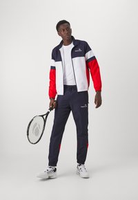 Track jacket with navy, red, and white panels, paired with navy pants featuring red accents, worn by a person holding a tennis racket.