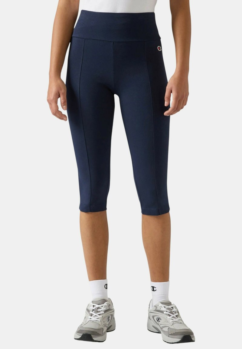 Femme portant des leggings courts bleu marine à taille haute, des chaussettes blanches avec un logo, et des baskets de sport grises sur un fond uni.