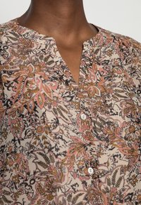 Chemise à boutons florale en tissu beige avec des motifs complexes roses, noirs et bruns, un col en V et des boutons blancs.