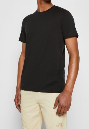 Man wearing a plain black short-sleeve t-shirt and light beige pants standing against a neutral background.