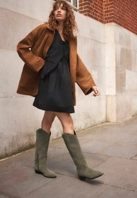 Chaqueta oversized de ante marrón sobre un vestido negro con cordón, combinada con botas de ante verdes hasta la rodilla con puntera afilada y cremalleras laterales.