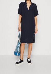 Robe courte bleu marine à manches courtes avec un décolleté en V, fentes latérales, associée à des baskets noires et un sac tissé bleu. Tissu doux, coupe décontractée.