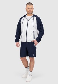 Giacca sportiva blu navy e bianca con cappuccio e zip, abbinata a pantaloni corti blu navy. Presenta loghi e accenti a contrasto. Sneakers bianche completano l'outfit.