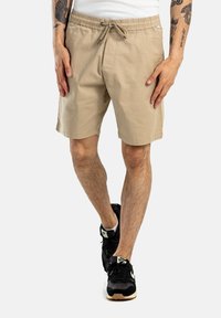 Pantalones cortos beige hechos de una tela ligera con cinturilla elástica y cordón. Cuenta con bolsillos laterales y un ajuste relajado. Se combinan con zapatillas negras.