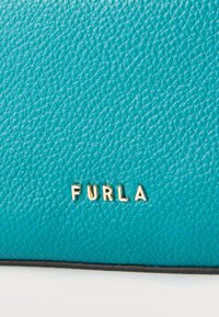 Γαλαζοπράσινη υφή δερμάτινης επιφάνειας με χρυσά μεταλλικά γράμματα που σχηματίζουν τη λέξη "FURLA" τοποθετημένα στο κέντρο κοντά στο κάτω άκρο.