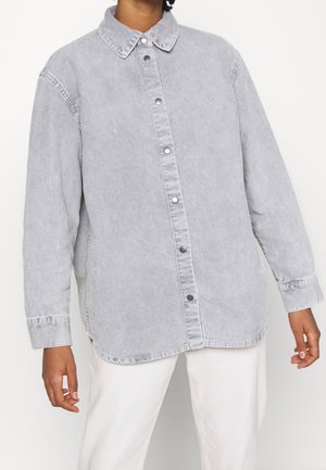 Personne portant une chemise boutonnée en velours côtelé gris clair à manches longues et un pantalon blanc sur un fond uni.