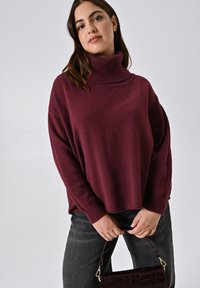 Burgunderfarbener Rollkragenpullover aus weichem Material mit geripptem Design. Kombiniert mit dunkelgrauen Jeans und einer Handtasche im Krokodil-Muster.