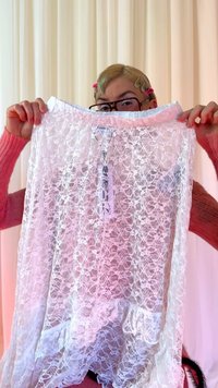 Άτομο με γυαλιά και κλιπ μαλλιών που κρατάει ένα διάφανο λευκό δαντελένιο ρούχο μπροστά από φόντο με φωτεινές κουρτίνες.