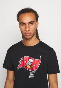 T-shirt en coton noir avec un graphique de drapeau des Buccaneers rouge représentant un crâne et des épées, avec un design classique à col rond et des manches courtes.