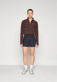 Ruda sportinė striukė su užtrauktuku, juodi šortai, balti kojinės ir sportbačiai. Striukėje yra du kišenės, o apranga pagaminta iš tamprios medžiagos.