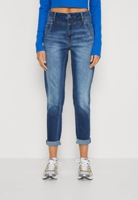 Person wearing rolled-up blue denim jeans, blue long-sleeve crop top, and white-gray sneakers, standing against white background.