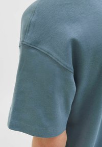 Manica corta di una t-shirt in cotone blu-grigio attenuato con cuciture visibili sulla spalla e sull'orlo della manica, su uno sfondo chiaro.