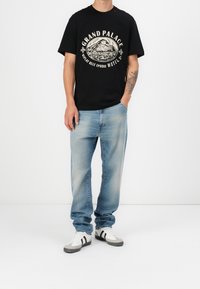 Schwarzes T-Shirt mit Berggrafik und dem Text "GRAND PALACE", kombiniert mit hellblauen Jeans und weißen Schuhen mit schwarzen Streifen.
