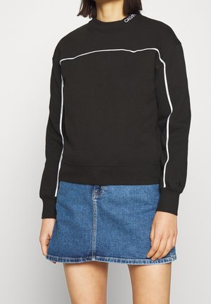 Personne portant un sweat-shirt noir à manches longues avec un passepoil blanc et une jupe en jean bleue sur un fond uni.