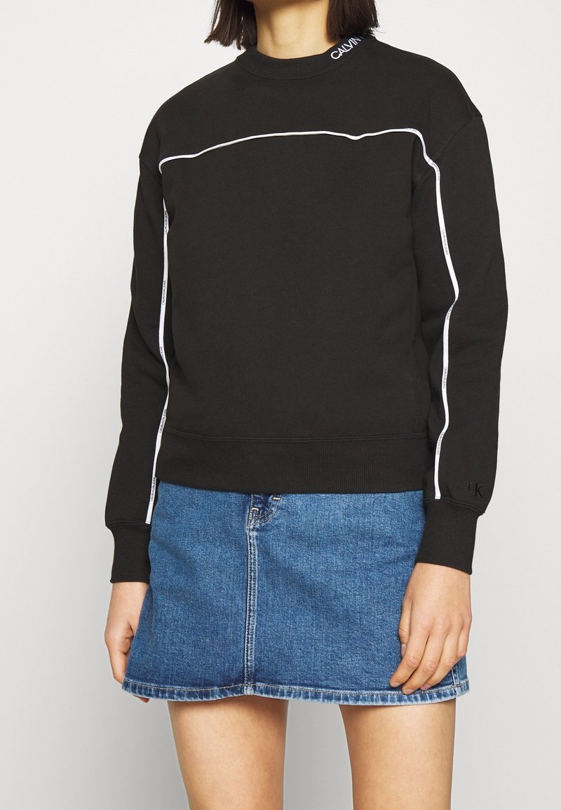 Personne portant un sweat-shirt noir à manches longues avec un passepoil blanc et une jupe en jean bleue sur un fond uni.