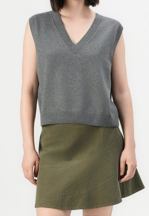 Femme portant un haut en maille gris sans manches à col en V et une jupe évasée vert olive sur un fond uni.