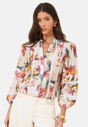 Vrouw die een doorschijnende blouse met lange mouwen draagt met een meerkleurige bloemenprint, kanten afwerking en crème hoge taillebroek, gecombineerd met gouden sieraden.