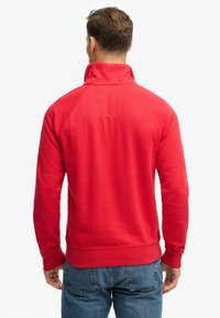 Sweat-shirt rouge à col montant avec une finition texturée. Caractérisé par des manches raglan et une coupe décontractée. Tissu en mélange de coton, détails minimaux.