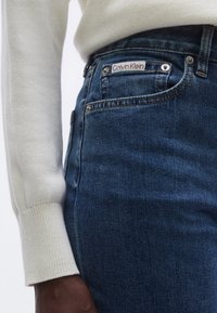 Tumesinised teksapüksid, mis istuvad tihedalt, tagataskul punase "Calvin Klein" sildiga ja hõbedaste detailidega. Pehme, tekstuuriga kangas.