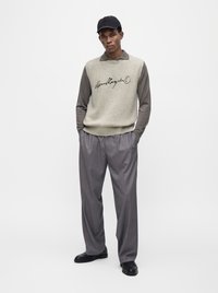 Άνδρας με μαύρο καπέλο, ανοιχτό γκρι αμάνικο πουλόβερ με κείμενο, γκρι μακριά πουκάμισα, φαρδιά γκρι παντελόνια και μαύρα παπούτσια στέκεται με τα χέρια στις τσέπες.
