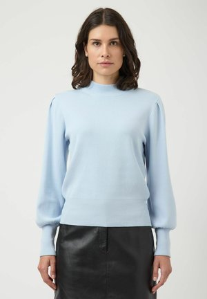 Kvinde med skulderlangt mørkt hår iført en lyseblå langærmet sweater og et sort lædernederdel, stående mod en ensfarvet baggrund.