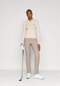 Egy nő áll, a jobb kezében tart egy golfütőt, bézs hosszúujjú pólót, világosbarna nadrágot, fehér kesztyűt és fehér sportcipőt visel.