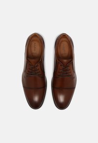Chaussures habillées en cuir brun avec une texture lisse, dotées d'un bout arrondi, de surpiqûres décoratives et d'un laçage. Nom de la marque visible à l'intérieur.