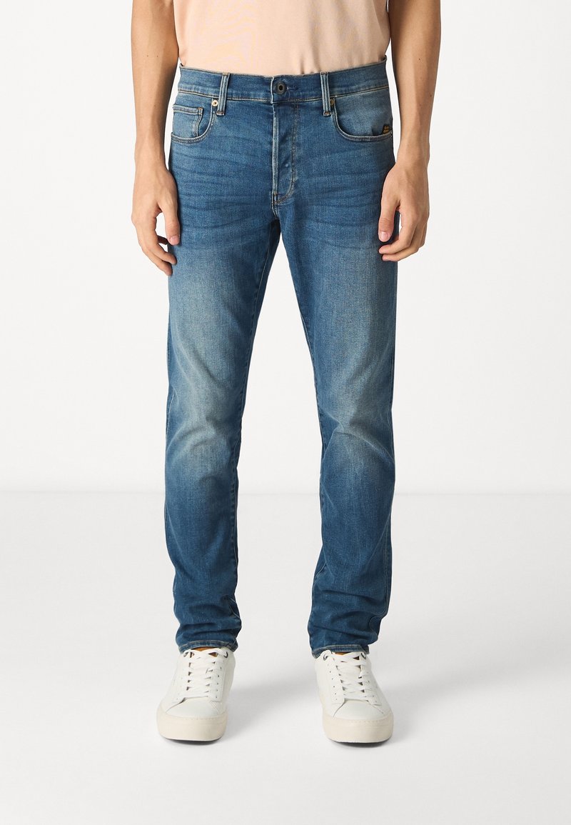 Blå denimjeans i slim fit. Har femfickorsdesign, subtil blekning och knappstängning. Bärs med vita sneakers.