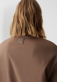 Bruine stoffen kleding met een gladde textuur, voorzien van een vierkante logo patch in een donkerdere bruintint op de achterkant van de halslijn.