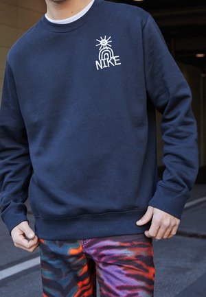 Osoba nosząca granatową bluzę z białym uśmiechniętym słońcem i tęczą nad słowem Nike, w zestawie z kolorowymi, wzorzystymi spodniami.