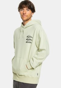Sudadera verde clara con bolsillo canguro, capucha ajustable con cordón y un estampado gráfico que presenta montañas en el lado izquierdo del pecho.