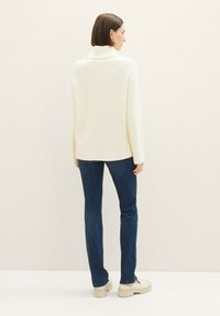 Chunky, kremfarget cardigan med høy krage, ribbestrikket tekstur og sidesliss. Kombinert med mørke jeans og beige plattformsneakers.