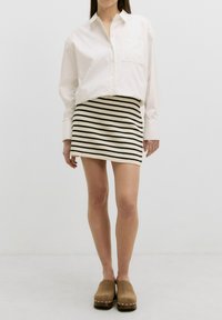 Chemise blanche à boutons avec manches longues, associée à une mini-jupe rayée noir et blanc. Des sabots marron complètent la tenue.