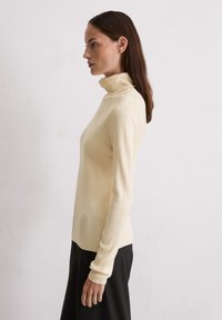 Haut à col roulé côtelé en beige clair avec des manches longues et une silhouette ajustée, associé à un vêtement noir en dessous de la taille.