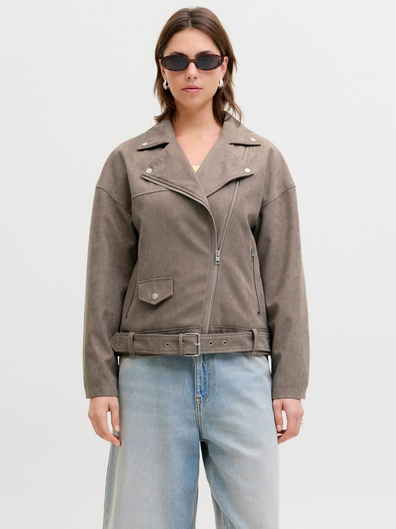 Vrouw met een bruine suède motorjack met riem, lichtgewassen wijde jeans, zilveren oorringen en donkere zonnebril.