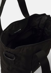 Saco tote de tecido preto com fecho zip, apresentando um bolso interior e logotipo impresso no interior. Inclui alças robustas e uma textura suave.
