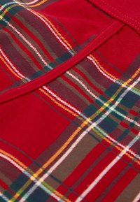Czerwona tkanina z wzorem tartanowym, na którym widoczne są zielone, żółte i brązowe linie. Gładka faktura, żywe, kontrastujące kolory oraz przeszywane brzegi.