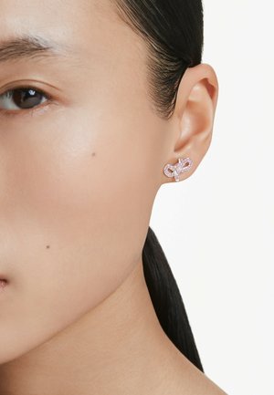 Rosa Edelsteinohrstecker im Schleifen-Design mit funkelnden Akzenten, in einem glänzenden Metall gefasst, getragen am Ohr eines Modells.