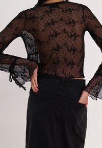 Personne portant un haut en dentelle florale noire transparente avec des manches longues et des poignets évasés, associé à un pantalon en denim noir taille haute.