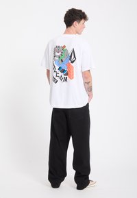 Jeune homme avec des tatouages portant un t-shirt blanc avec une illustration de dessin animé colorée et un pantalon noir, debout devant un fond uni.