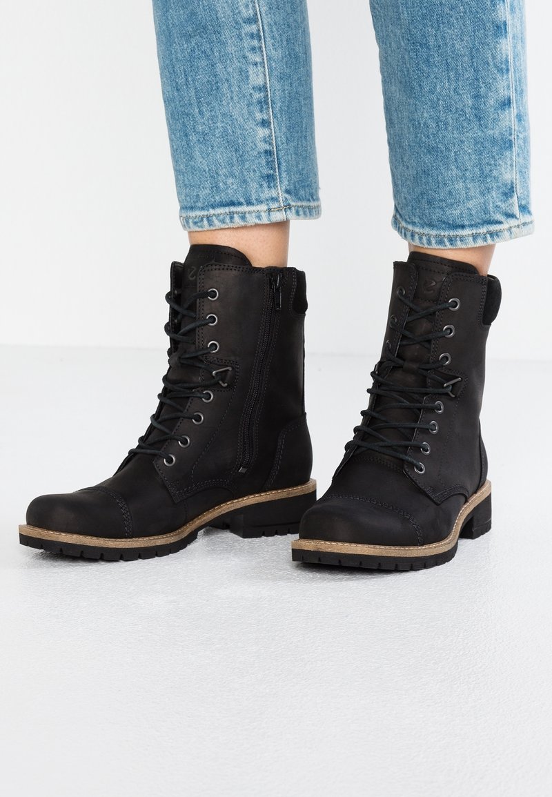 Bottines noires à lacets avec fermetures éclair latérales portées avec un jean bleu coupé au-dessus de la cheville sur une surface blanche.