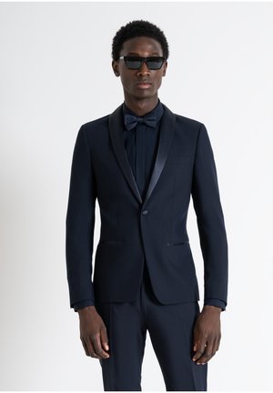 Completo blu navy scuro con rever in satin, papillon coordinato e un design aderente. Indossato con occhiali da sole scuri. Tessuto liscio e vestibilità su misura.