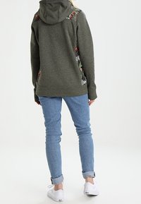 Sudadera verde oliva con un acabado texturizado, adornada con patrones florales en los hombros, combinada con jeans de un azul claro y zapatillas blancas.