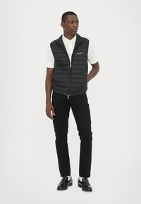 Sort dunvest med høj krave, lynlåslukning, båret over en hvid polo skjorte, parret med sorte bukser og sko. Simpelt design, minimale detaljer.