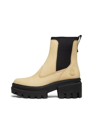 Beige ankelstøvle med chunky sort slidstærk sål, sorte elastiske sidepaneler og trækkeøje bagpå, med en lille præget logo nær hælen.
