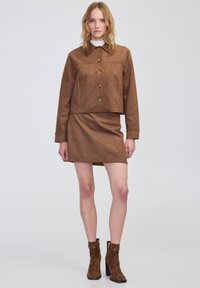 Chaqueta marrón corta con botones, falda corta marrón a juego y botas de tobillo de ante marrón. Textura lisa, diseño minimalista. Fondo liso.