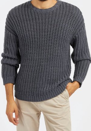 Mand iført en stor mørkegrå strikket sweater med rund hals og beige bukser med den ene hånd i lommen mod en ensfarvet baggrund.